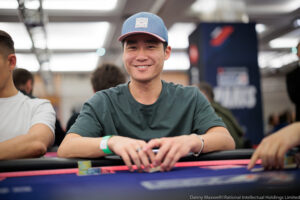 Smiling young man in a blue cap and green shirt sits at a poker table with chips in front of him.
